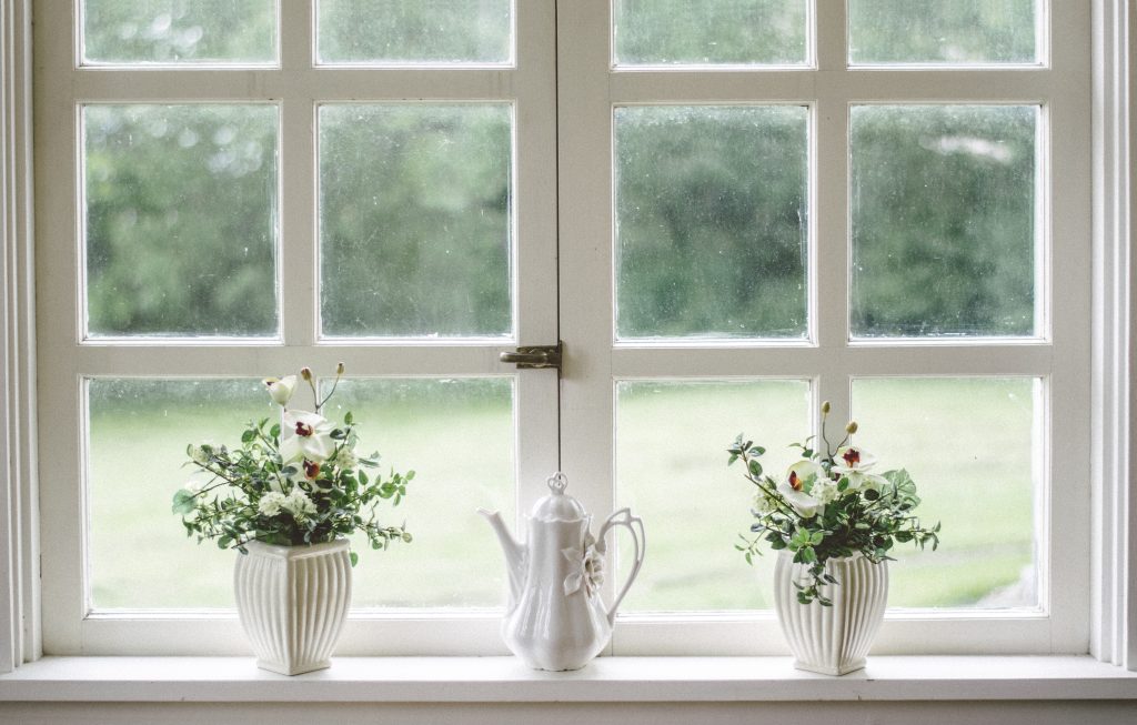 
A white-framed window with multiple panes, displaying a serene view of greenery outside. On the windowsill, there are two white vases with delicate flowers and a white teapot, creating a charming and peaceful scene.