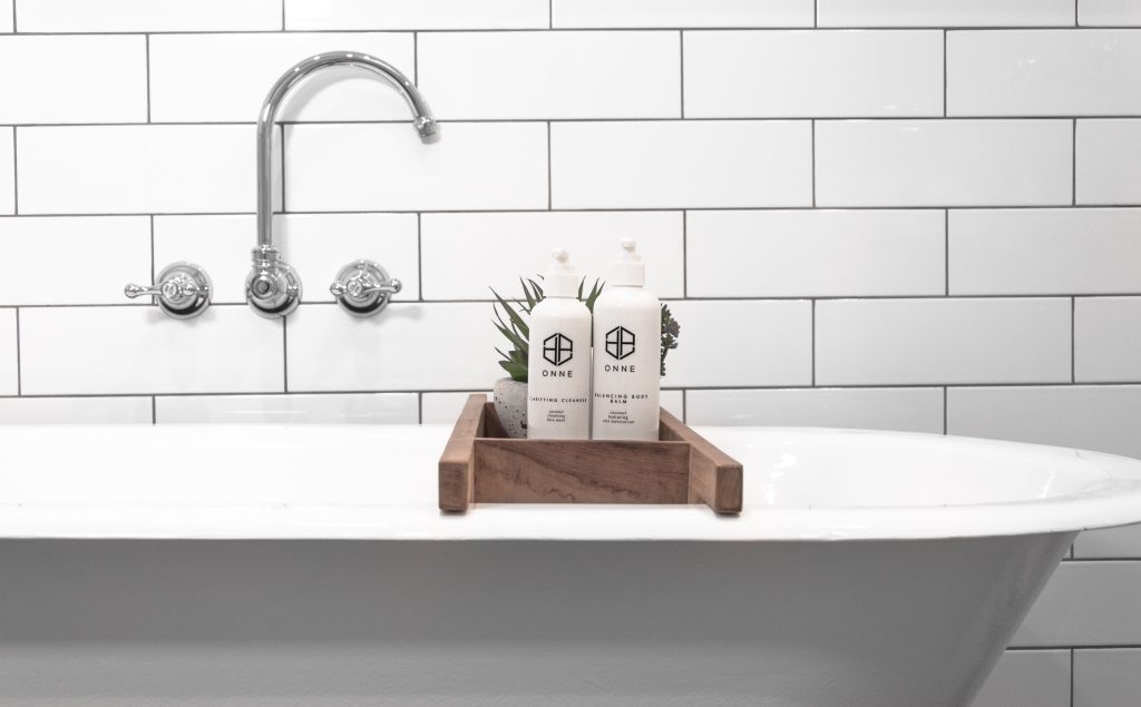White bathtub with a wooden tray holding two bottles of ONNE skincare products and a plant, set against a white tiled wall with chrome fixtures