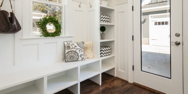 A bright and tidy entryway featuring built-in white shelves and cubbies. The area is decorated with patterned cushions, a fluffy throw, and a wreath hanging in the window. The glass door offers a view of the driveway and garage outside