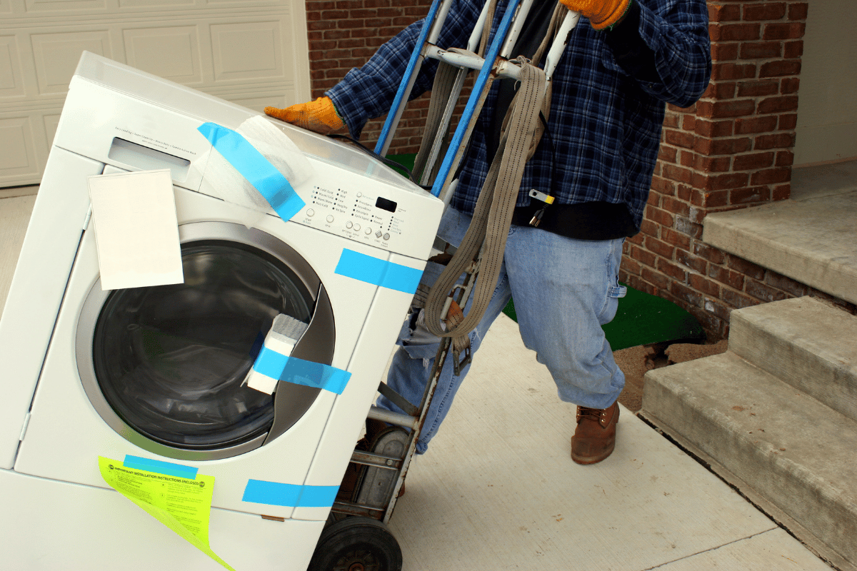 a removalist moving a washing machine. the removalist uses a trolley, a moving belt and has the laundry machine taped up
