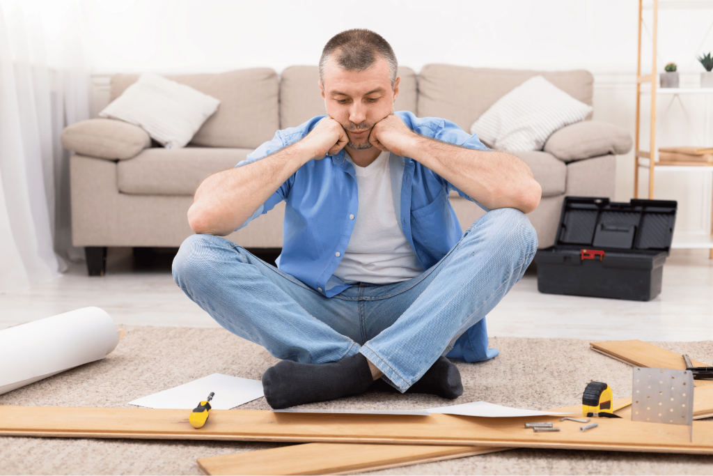 A man sits cross-legged on the floor, looking frustrated and resting his head on his fists. In front of him are tools and assembly instructions, indicating he is struggling with a DIY project. In the background, a couch and a toolbox can be seen.
