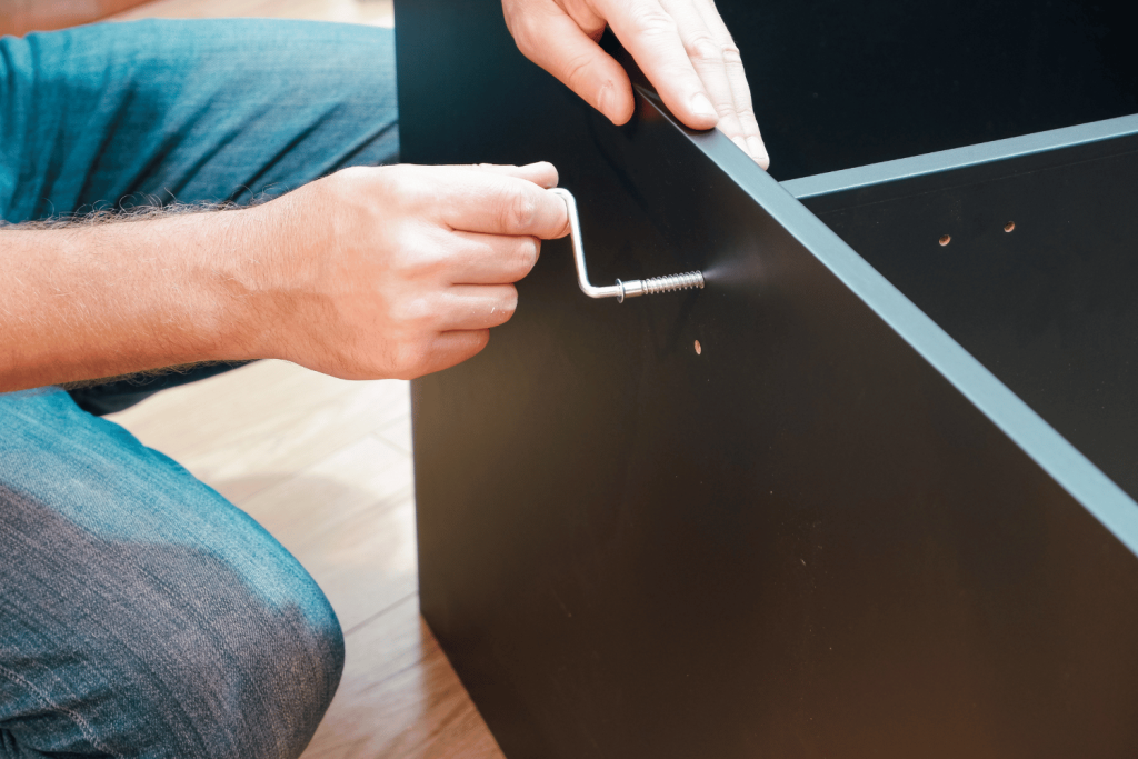 
A close-up of a person assembling furniture using an Allen wrench. The person's hands are visible, tightening a screw on a black piece of furniture.