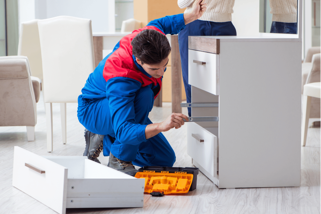 
A man in a blue and red uniform is assembling a white chest of drawers. He is using a tool to secure the drawer slides while a toolbox and a removed drawer are on the floor nearby. Another person is standing beside him, indicating teamwork in the assembly project.