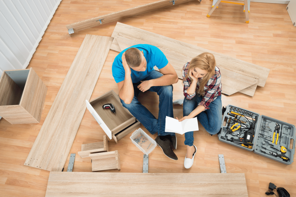 A man and a woman sit on the floor surrounded by unassembled furniture pieces and tools. The man looks frustrated with his hand on his head, while the woman reads the assembly instructions. A toolbox and various parts are scattered around them, indicating a furniture assembly project.