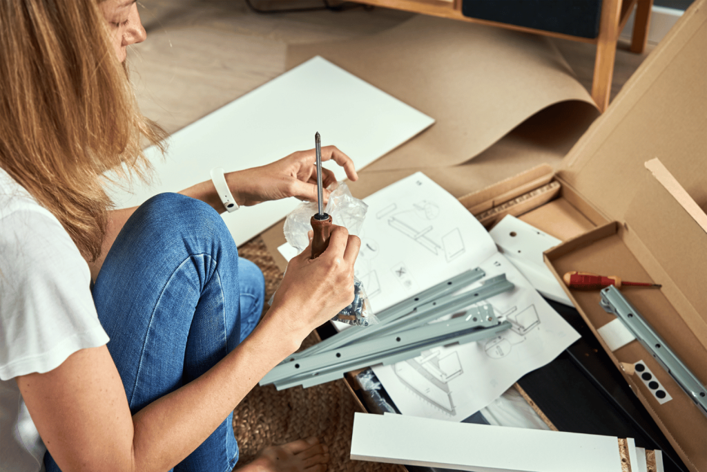 
A woman sits on the floor assembling furniture. She is holding a screw and using a screwdriver, surrounded by instruction manuals, metal rails, and various parts